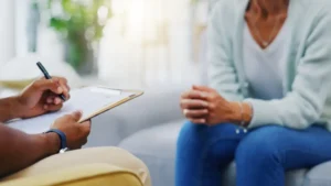 Person holding a clipboard talking with another seated person in a clinical setting